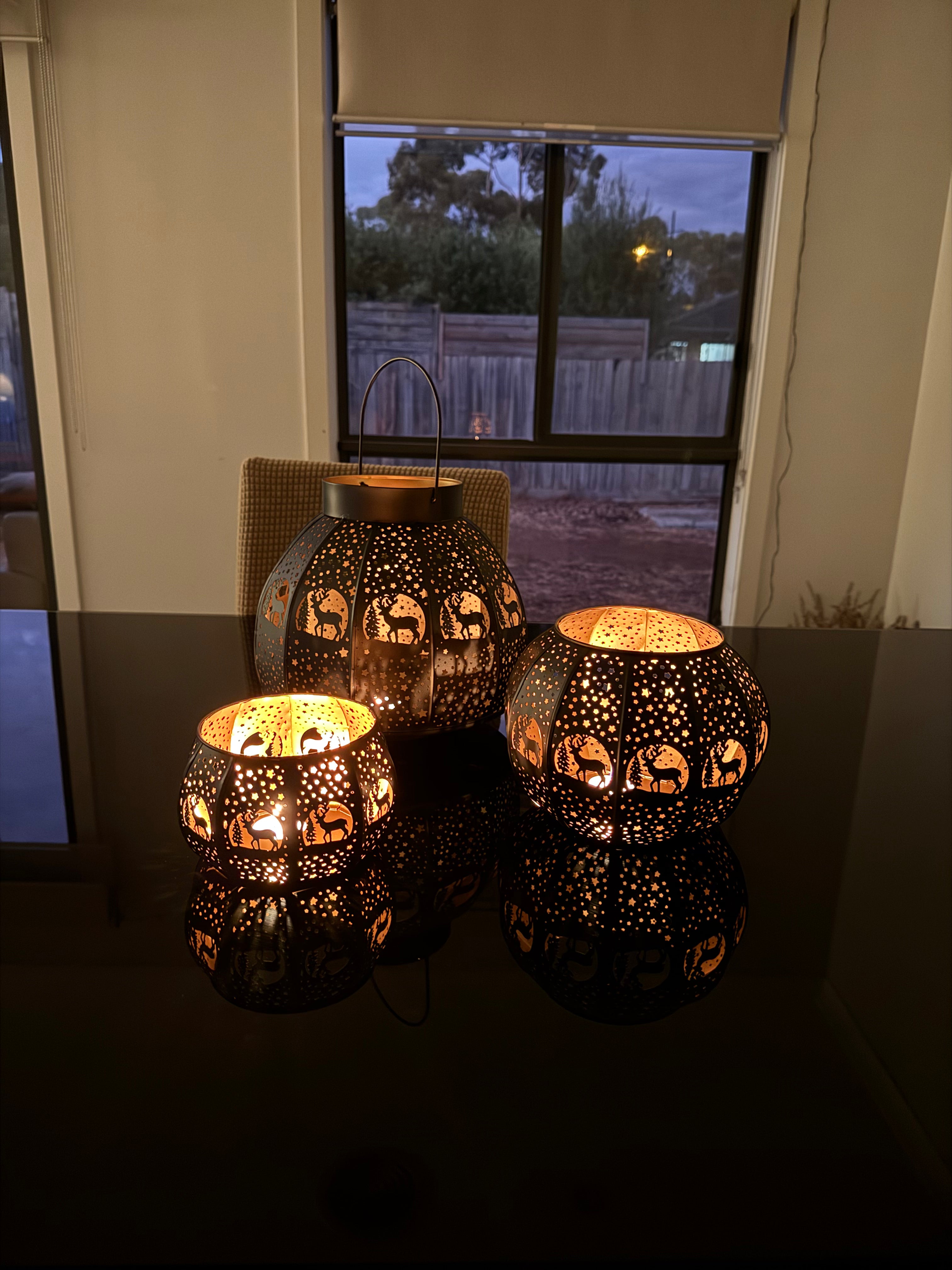 Three brass candle lamps with intricate etchings, displaying a warm glow, placed on a surface near a window.